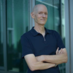 Author Lee Schneider wearing a blue shirt, standing with arms folded in front of a green building.
